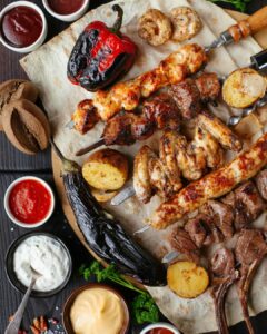 Skewered and barbecued meats along side dipping sauces, flame roasted vegetables on a wooden table. 