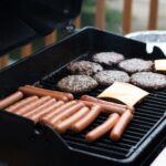 Hot dogs and hamburgers lay spread across an open barbecue. Cheese is being applied to some of the burger paddies.