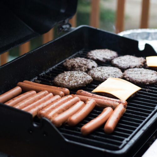 Hot dogs and hamburgers lay spread across an open barbecue. Cheese is being applied to some of the burger paddies.