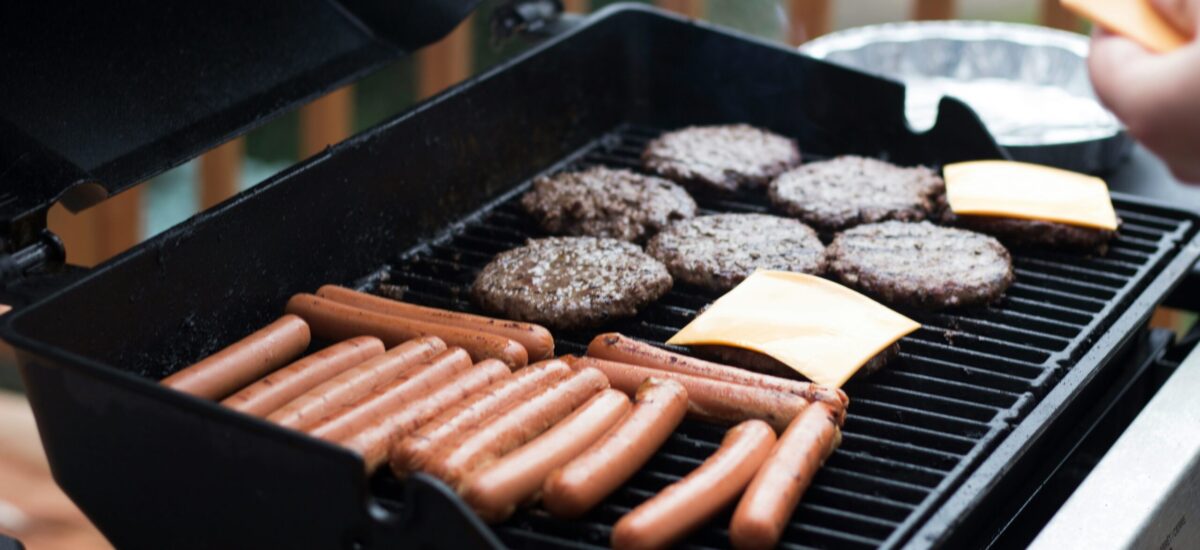 Hot dogs and hamburgers lay spread across an open barbecue. Cheese is being applied to some of the burger paddies.