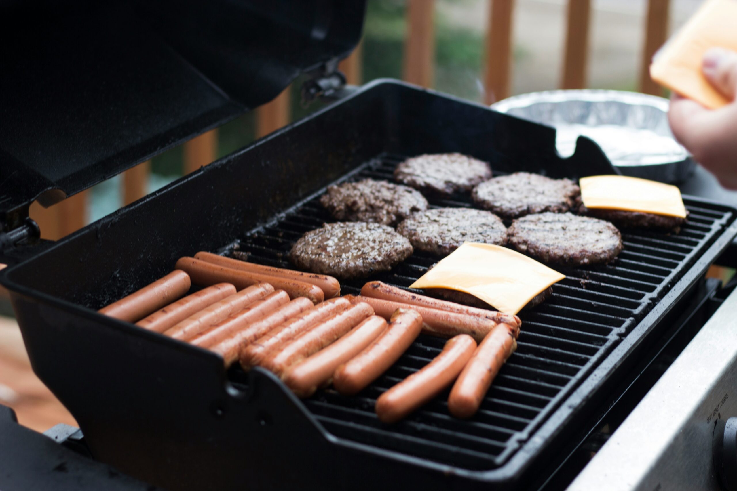 Hot dogs and hamburgers lay spread across an open barbecue. Cheese is being applied to some of the burger paddies.